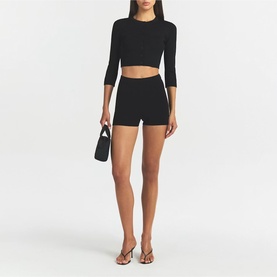 Model wearing Skims black shorts with matching black crop top, styled with clear heeled mules and a small black bag