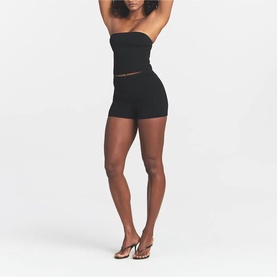 Model wearing Skims black strapless bandeau top with sheer waist detail, styled with black shorts and clear heeled mules