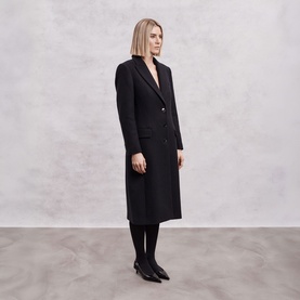 Model wearing The Landskein black long coat, styled with black tights and black pointed-toe flats, side profile view.