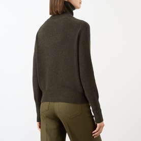 Model wearing Victoria Beckham Marie Turtleneck Sweater in dark green wool, rear view, with olive trousers. Ribbed trims and straight hem visible.