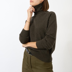 Model wearing Victoria Beckham Marie Turtleneck Sweater in dark green wool with ribbed trims, styled with an olive mini skirt and gold bracelet.