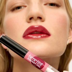 Model wearing Benefit Benetint Pocket Pal Lip Gloss in Original Rose, holding the tube, lips shown glossy and deeply pigmented pink-red.
