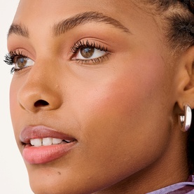 Model wearing Benefit You've Got Aura Summer Essentials Gift Set; dewy skin, rosy blush, fluttery lashes, nude lip, silver hoop earring.
