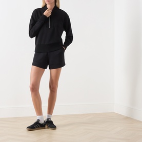 Model wearing Varley black half-zip sweat top with matching black shorts and grey trainers with white socks.