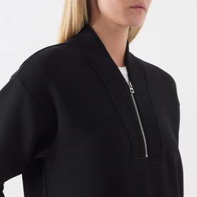 Model wearing Varley black sweat top, close-up showing half-zip neckline with ribbed collar detail and white underlayer visible.