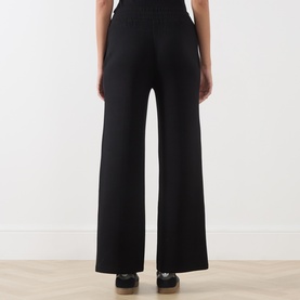 Model wearing Varley black wide-leg sweatpants, rear view, shown with black trainers on a light wood floor.