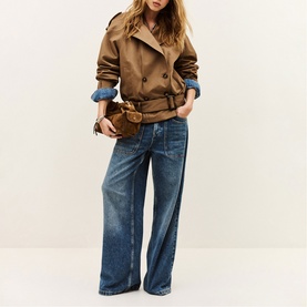 Model wearing Bash brown belted short trench jacket with denim cuffs, styled with wide-leg blue jeans, carrying a brown clutch bag