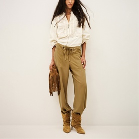 Model wearing Bash yellow relaxed-fit trousers with drawstring waist, styled with white zip blouse and tan suede boots, carrying fringed bag