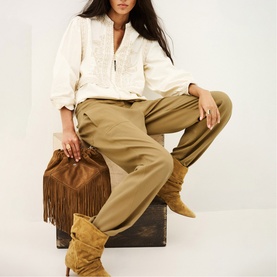 Model wearing Bash yellow trousers with white embroidered blouse, styled with tan suede knee-high boots and brown fringe bag