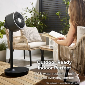 Dimplex black pedestal fan on outdoor decking beside rattan chairs, with model reading in background. UV and water resistant design.