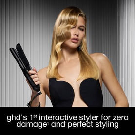 Model with GHD hair straightener styling long blonde hair, holding black flat iron tool against a striped background