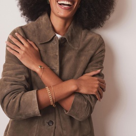 Model with Mint Velvet gold chain bracelets stacked on wrist, styled with an olive suede shirt jacket.