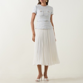Model wearing Self-Portrait blue tweed-effect top and white pleated midi skirt dress with gold button details, styled with white heeled mules.