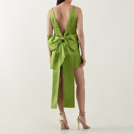 Model wearing Self-Portrait green dress with deep V back, oversized bow detail, thigh-high split, styled with gold strappy heels.