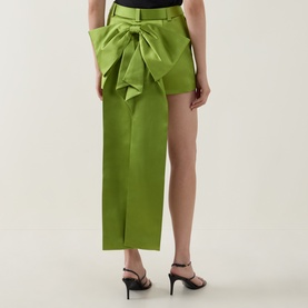 Model wearing Self-Portrait green satin midi skirt with oversized bow detail at waist, styled with black strappy heeled sandals.