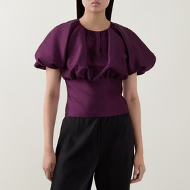 Model wearing Self-Portrait purple puff-sleeve top with gathered neckline and fitted waist, styled with black trousers.