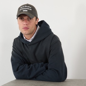 Model with Acne Logo Baseball Cap in faded black, embroidered Stockholm 1996 text, peaked brim. Styled with dark navy hoodie.