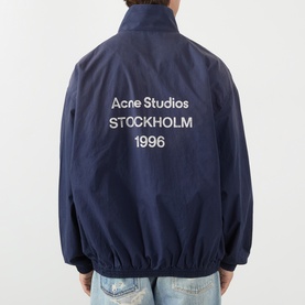 Model wearing Acne navy cotton casual jacket, back view showing white Acne Studios Stockholm 1996 logo print, styled with light-wash jeans.
