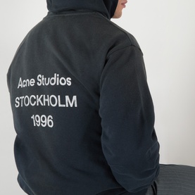 Model wearing Acne black cotton hoodie sweatshirt, rear view showing Acne Studios Stockholm 1996 logo print in white.