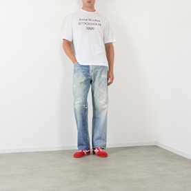 Model wearing Acne 1996 Logo T-Shirt in dusty white with light wash distressed jeans and red sneakers.