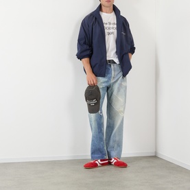 Model wearing Acne 1996 Logo T-Shirt in dusty white with navy jacket, light wash jeans, red trainers, holding dark cap.