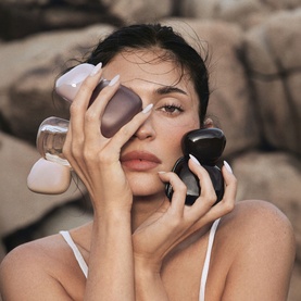 Model with Kylie Fragrance EDPs, holding multiple black and nude bottle caps close to face, rocky outdoor backdrop