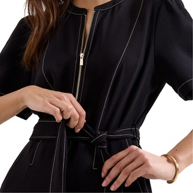 Model wearing Phase Eight black dress with contrast stitching, zip front, and tie waist belt; gold zip and bracelet visible.