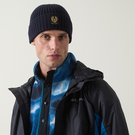 Model with Belstaff Watch Logo Beanie Hat in Dark Ink, ribbed-knit wool with logo patch, styled with navy jacket and blue scarf
