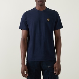 Model wearing Belstaff Phoenix Logo T-Shirt in Dark Ink, crew neck, short sleeves, gold embroidered chest logo, styled with dark jeans.