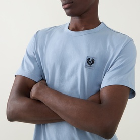 Model wearing Belstaff Phoenix Logo T-Shirt in Mist Blue, crew neck, short sleeves, small chest logo, arms crossed.
