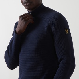 Model wearing Belstaff Dark Ink cotton half-zip knit, featuring textured stitch detail and gold logo badge on sleeve.