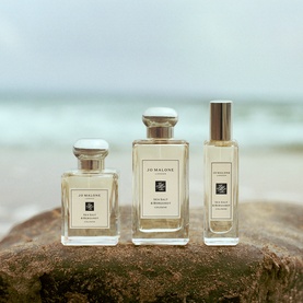 Jo Malone London cologne trio in clear glass bottles with silver caps, displayed on driftwood with ocean backdrop