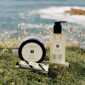 Jo Malone Bath & Body set styled outdoors on coastal grass, featuring a pump bottle, body cream jar, and travel-size tube