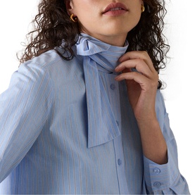 Model wearing Whistles blue striped shirt with self-tie neck bow, styled with gold hoop earrings. Close-up detail shot.