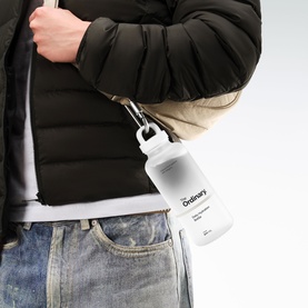 Model with The Ordinary Daily Hydration Bottle, white with branded label, clipped via carabiner, styled with dark puffer jacket and jeans