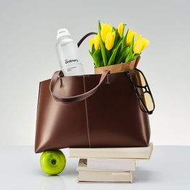 The Ordinary Daily Hydration Bottle in white, shown upright inside a brown leather tote bag styled with yellow tulips and sunglasses.