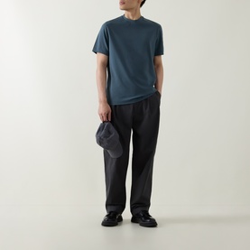Model wearing Belstaff Alloy Logo T-Shirt in petrol, styled with black wide-leg trousers and black loafers, holding a grey cap.