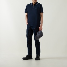 Model wearing Belstaff Alloy Logo Tipped Zip Polo Shirt in Dark Ink, styled with navy cargo trousers and black boots, holding a grey cap.