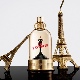 Jean Paul Gaultier La Favorite Eau de Parfum in gold bell jar bottle, flanked by two miniature Eiffel Tower ornaments on dark surface