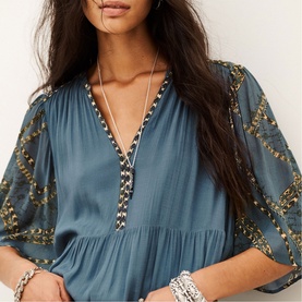 Model wearing Bash blue V-neck blouse with gold embroidered sleeves, styled with layered necklace and bracelets.