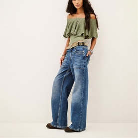 Model wearing Bash green off-shoulder ruffled top, styled with wide-leg blue jeans, brown belt and dark loafers