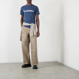 Model wearing Acne Stained Twill Trousers in camel beige, wide-leg fit with upturned hems, styled with a blue logo tee and tied shirt.