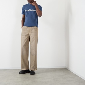 Model wearing Acne Stained Twill Trousers in camel beige, wide-leg with upturned hems, styled with a blue logo tee and black shoes.