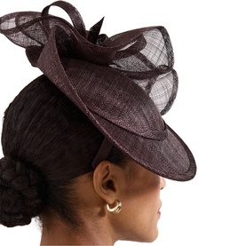 Model wearing Phase Eight brown sinamay fascinator with curved brim and looped bow detail, styled with gold hoop earrings