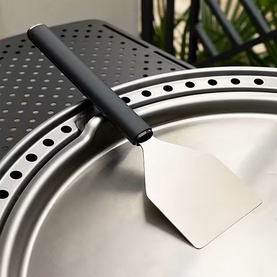 Stainless steel pizza peel with black handle resting on a perforated round grill lid