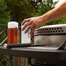 Solo Stove portable grill on outdoor surface beside Solo fuel bottle, hand reaching toward lid, green foliage in background