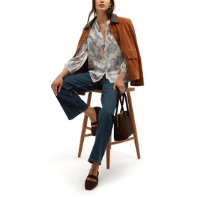 Model wearing Hobbs blue multi printed blouse and jeans, styled with a tan jacket, brown bag and mule sandals, seated on wooden stool