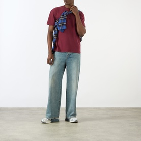 Model wearing Pellador maroon cotton T-shirt with light-wash wide-leg jeans and blue plaid shirt tied over shoulder.