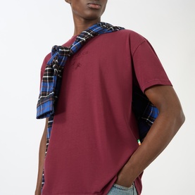 Model wearing Pellador maroon cotton T-shirt, styled with a blue tartan shirt tied over the shoulder.