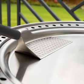 Close-up of a silver BBQ grill lid with ventilation holes and textured handle detail, garden visible in background